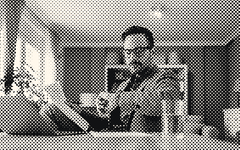 Un homme à lunettes consulte sa montre, un document à la main, assis à une table devant un ordinateur portable.