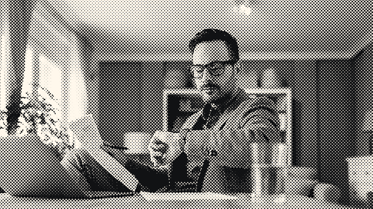 Un homme à lunettes consulte sa montre, un document à la main, assis à une table devant un ordinateur portable.