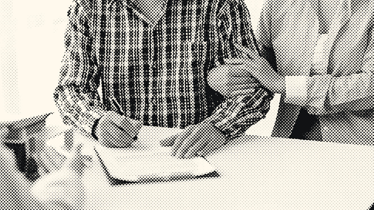 A man in a plaid shirt signs a form on a clipboard while a woman holds his arm; a small house model sits on the desk