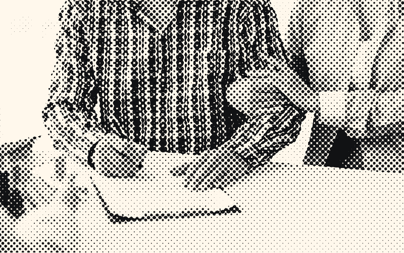 Un homme en chemise à carreaux signe un formulaire sur une planchette à pince pendant qu’une femme lui tient le bras; une petite maquette de maison est posée sur le bureau.